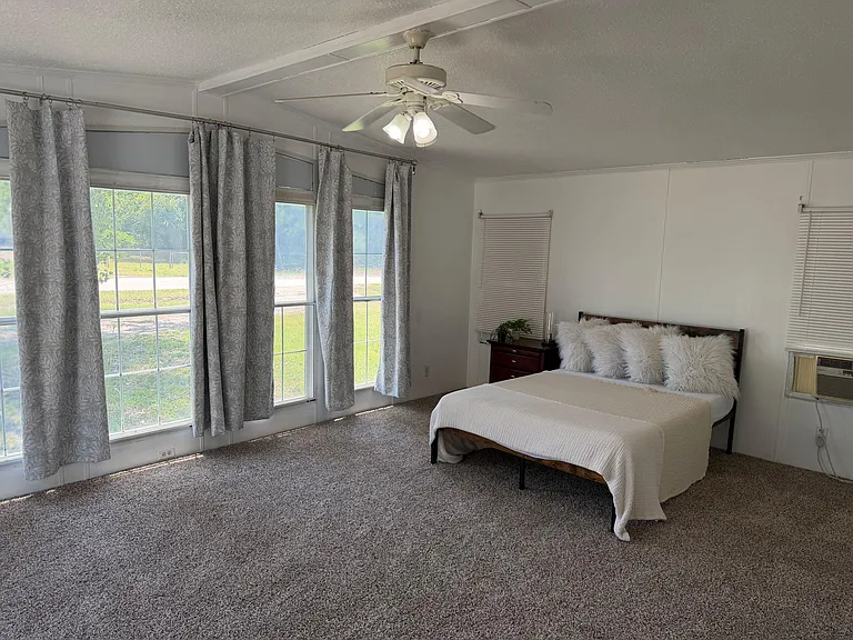 Spacious bedroom with large windows, curtain panels, ceiling fan, and carpeted floorsideal for a restful setup.