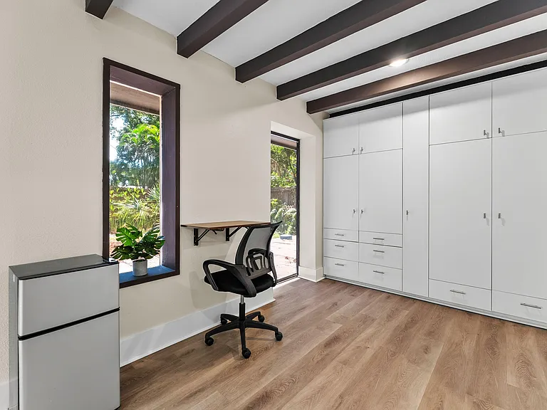 Room features exposed ceiling beams, large windows, built-in wardrobe storage, and a compact work area.