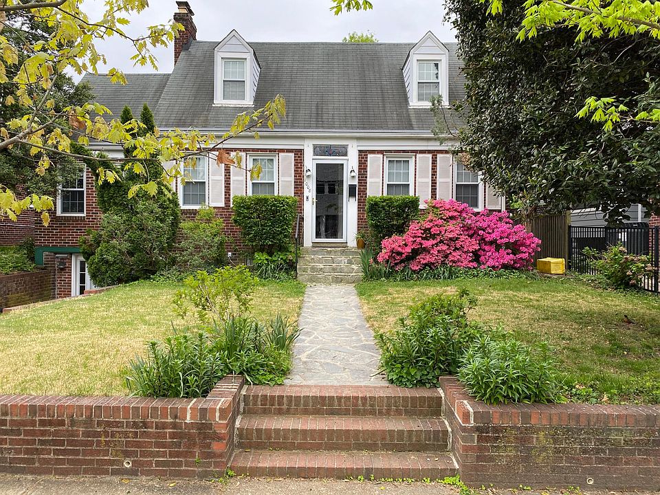 Front view of house, featuring mature plantings, brick retaining wall, and both off-street driveway parking for one care and permitted street parking.