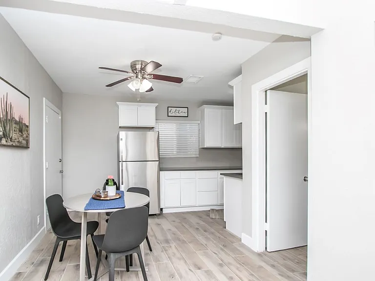 Detached casita dining and kitchen area at, Tempe rental home. Features a round dining table with modern black chairs, a stainless steel refrigerator, white shaker cabinets, wood-plank flooring, and desert wall art. Open layout for a top Tempe, AZ stay.