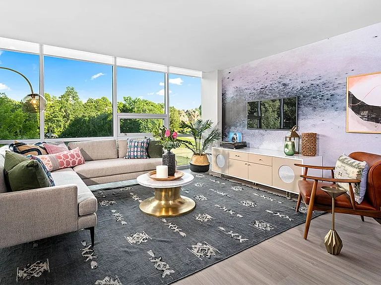 A living room with a grey couch, a round gold coffee table, and a large window with a view of trees.