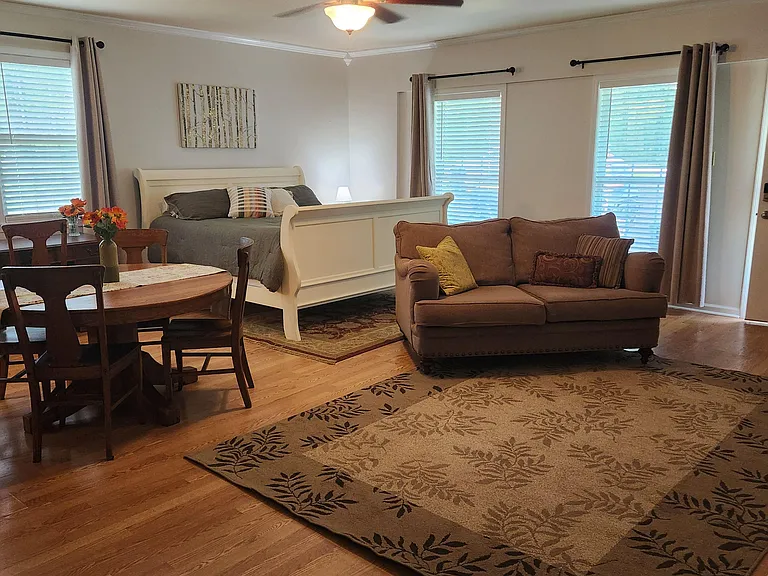 Spacious studio-style room with a defined sleeping area, dining table, and seating, plus multiple windows and ceiling fan.