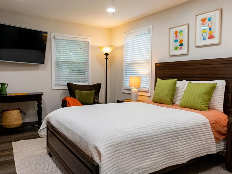 Furnished bedroom with a queen bed, nightstand lighting, desk area, and two windows with blinds for privacy.