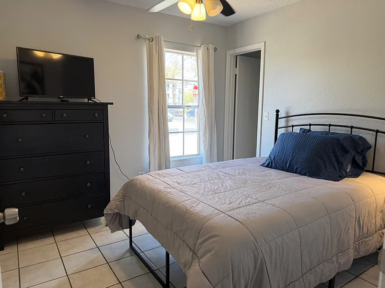 Furnished bedroom with a full bed, dresser with TV, and a window with curtains for natural light.