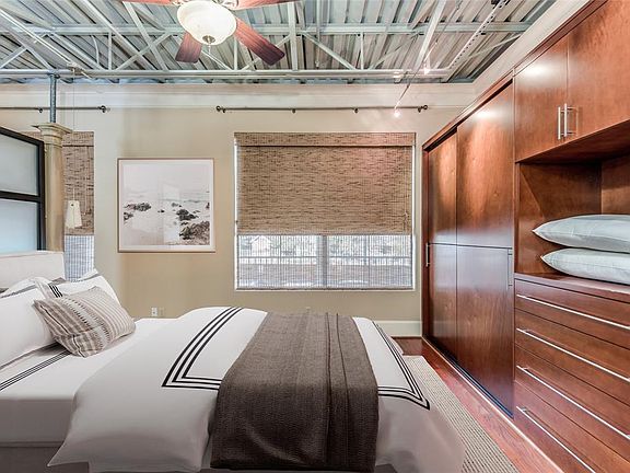 This virtually staged photo highlights the warmness of the primary bedroom area with high exposed ceilings, bamboo shades and custom built-in closet