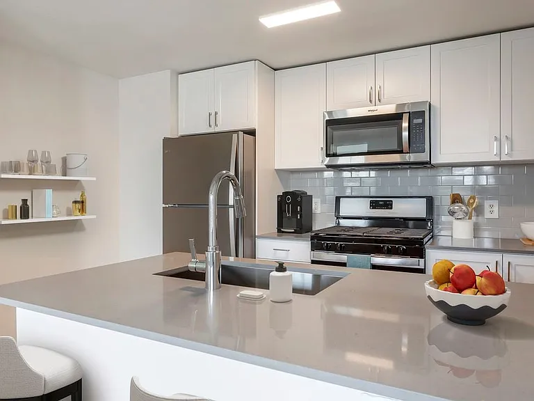 Finish Package II kitchen with white cabinetry, grey quartz countertops, extended hard surface flooring in select units, stainless steel appliances, and tile backsplash