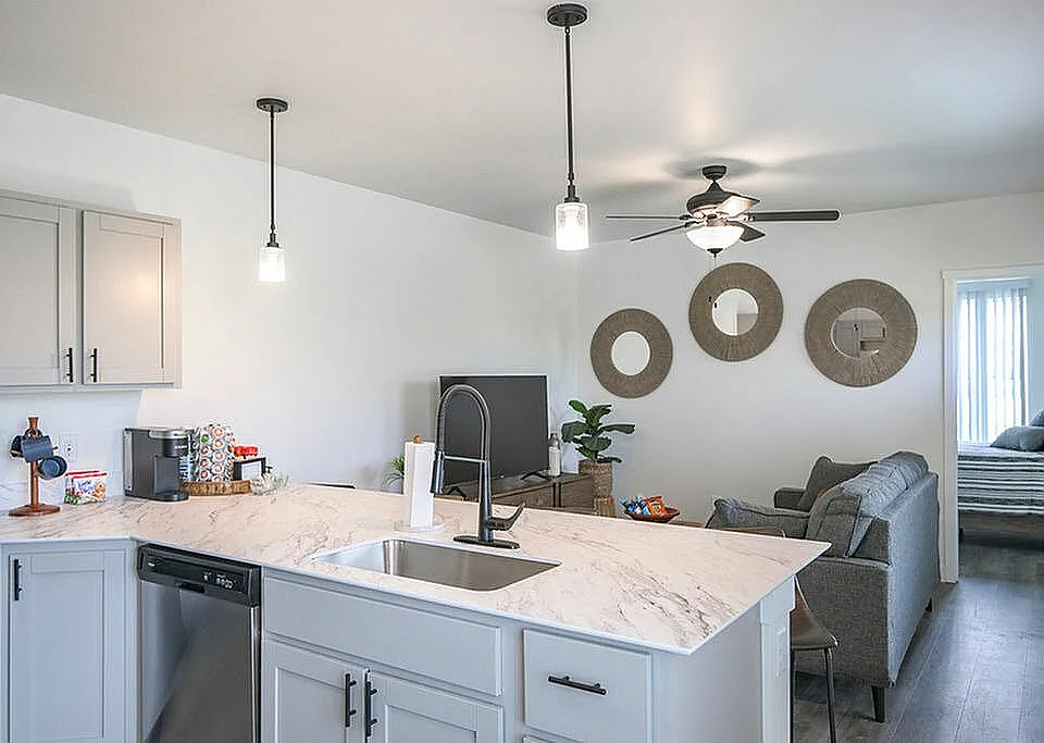 Elegant modern living space featuring open-concept kitchen with marble countertops, cozy living area, stylish decor, and view into a bright bedroom.