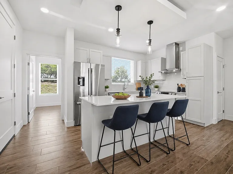 Contemporary kitchen featuring a large island with quartz countertops, stainless steel appliances, light gray cabinetry, and wood-look tile flooring; the door on the left opens to a private office.