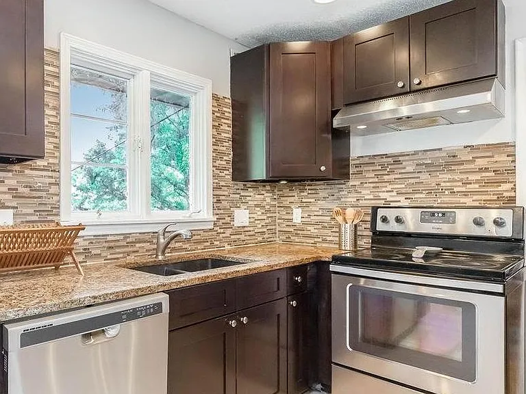 Efficient L-shaped kitchen with granite-look counters, tile backsplash, stainless range and dishwasher, plus a window over th