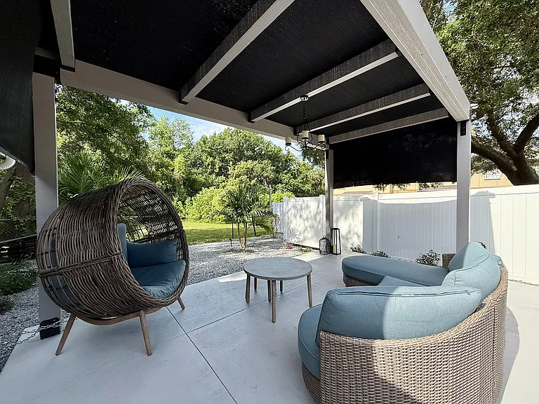 Charming patio with modern wicker furniture, featuring a cozy hanging chair and stylish seating, surrounded by lush greenery and shaded by an elegant canopy.