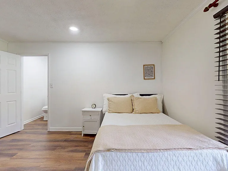 Comfortable bedroom with wood-style flooring, a large window with blinds, and space for a nightstand and storage.