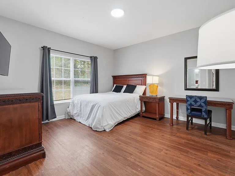 Spacious furnished bedroom with a queen bed, hardwood floors, TV, and a dedicated desk area beside a large window.