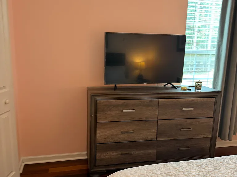 Bedroom with hardwood floors, window with blinds, and a dresser topped with a TV for convenient in-room storage.
