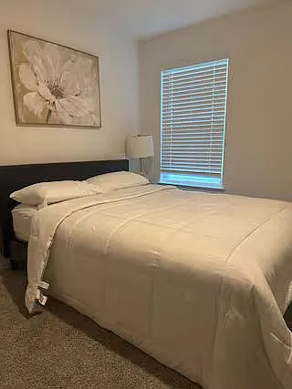 Comfortable bedroom with a full bed, soft carpet, bedside lighting, and a window with blinds for privacy.