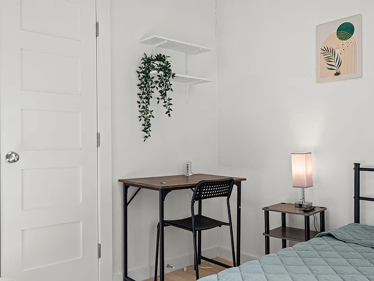 Bedroom includes a dedicated desk workspace, bedside table with lamp, and wall shelving for added storage and display.