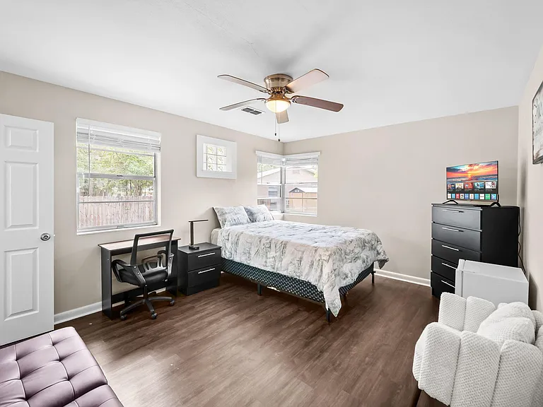 Spacious bedroom with two windows, ceiling fan, work desk, and dresser with TVideal for comfortable daily living.