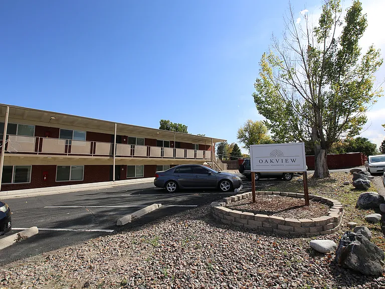 Oakview Apartments showcases a modern two-story building with ample parking under a clear blue sky, surrounded by lush trees and well-maintained landscaping, offering a serene living environment.