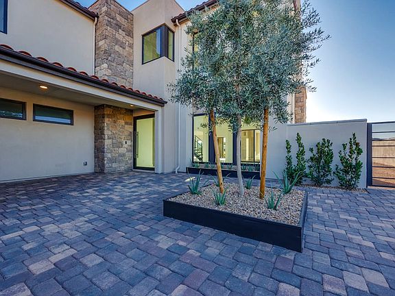A modern, two-story house with a paved courtyard in the foreground, featuring a large olive tree and
