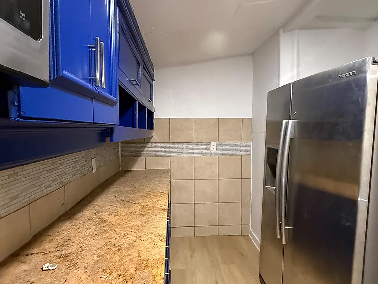 Compact kitchen with ample cabinet storage, tile backsplash, generous counter space, and a full-size stainless refrigerator.