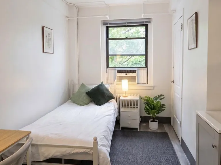 Efficient single bedroom with a twin bed, desk area, nightstand, and window A/C, plus a large window with leafy views.