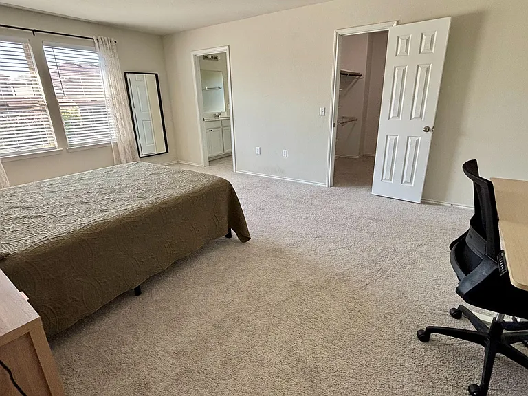 Spacious carpeted bedroom with large windows, a work desk, and a walk-in closet, plus direct access to a vanity area.