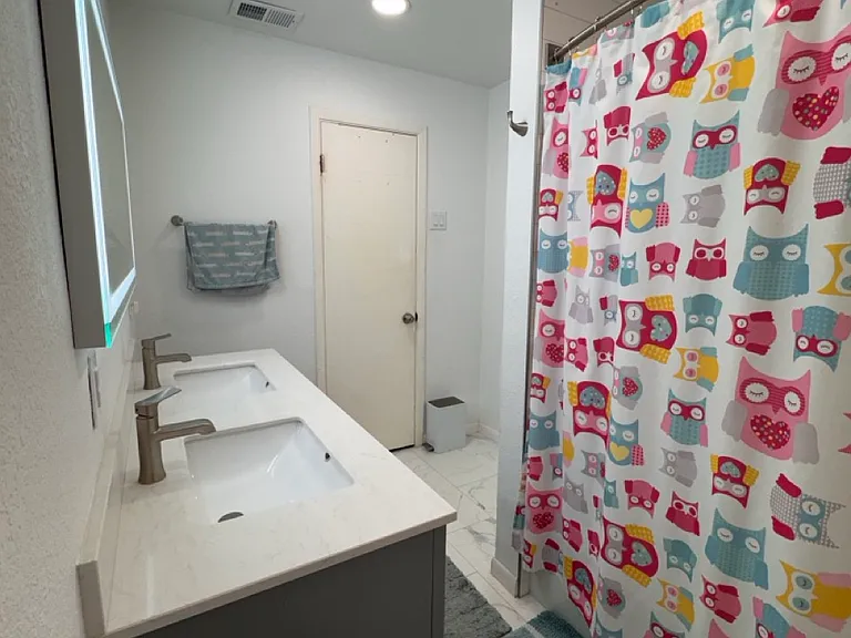The fully renovated second bathroom features a spacious double-sink vanity, an integrated LED-backlit mirror, beautiful marble-look tile, and a premium Kohler soaking tub with a full shower setup.
