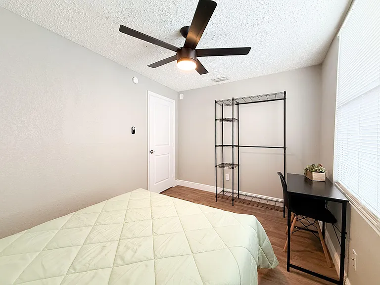 Private bedroom with ceiling fan, wood-style flooring, and window blinds, plus a desk area and open wardrobe shelving.