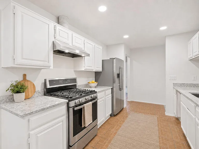 Galley kitchen with ample white cabinetry, stone-look counters, and stainless steel range hood, stove, and refrigerator.