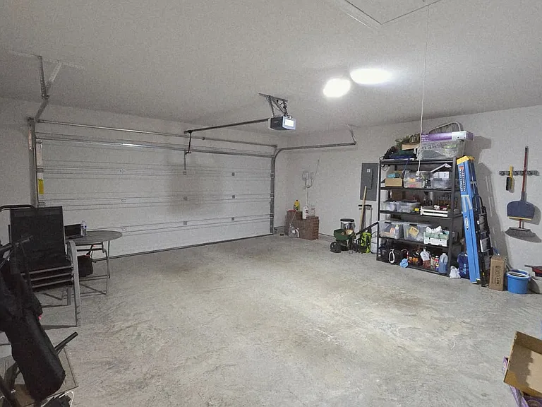 Bright and spacious two-car garage with a clean concrete floor and high ceiling. The space features a wide automatic garage door, strong overhead lighting, and a storage shelving area.