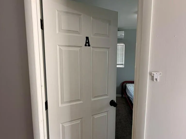 White interior door opening to a carpeted bedroom with a window and bed frame visible.