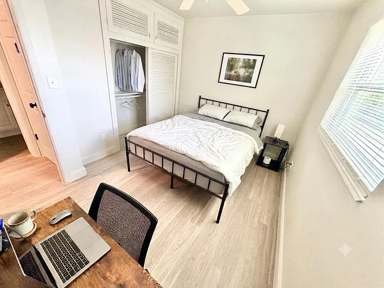 Furnished bedroom with a queen bed, side table, and built-in closet storage, plus a desk area and window blinds.