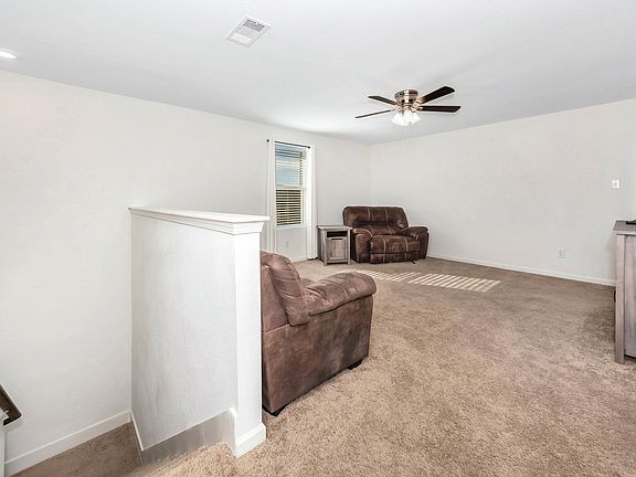 This spacious upstairs loft features neutral carpeting, crisp white walls, and a central ceiling fan. Furnished with plush brown recliners and a large television on a sleek stand, the area provides a functional and comfortable secondary living space.