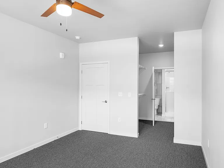 Contemporary room with sleek ceiling fan, grey carpeting, and access to a modern bathroom featuring clean lines and minimalist design. Contemporary room with sleek ceiling fan, grey carpeting, and access to a modern bathroom featuring clean lines and mini