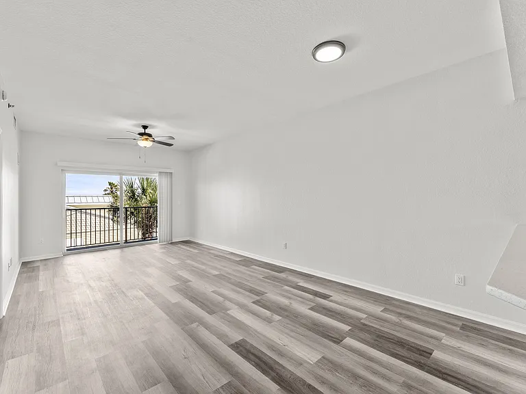 Modern living room with sleek wooden flooring, bright natural light, and a ceiling fan, opening to a balcony with a scenic view.