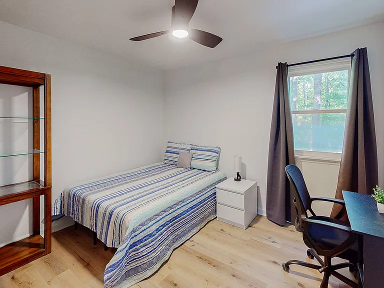 Furnished bedroom with a ceiling fan, bedside table, and dedicated desk space, plus a window with curtain panels.