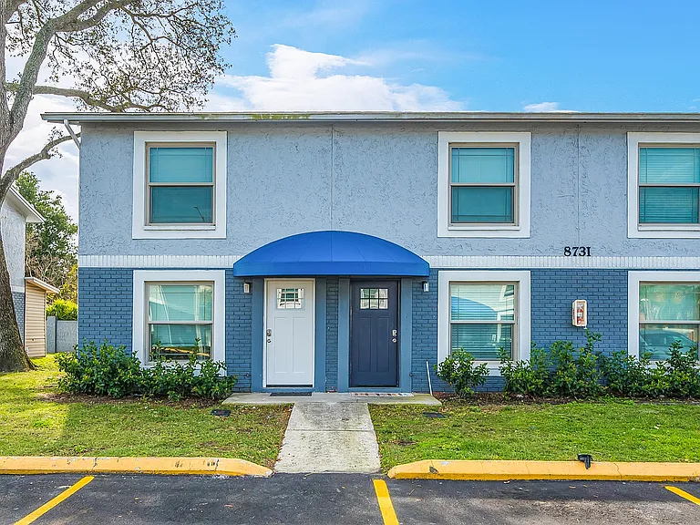 Charming two-story blue townhouse with lush landscaping, featuring large windows and an inviting entrance.