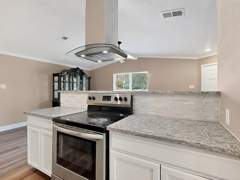 Kitchen with stainless steel electric range, white cabinetry, backsplash, ornamental molding, and island exhaust hood