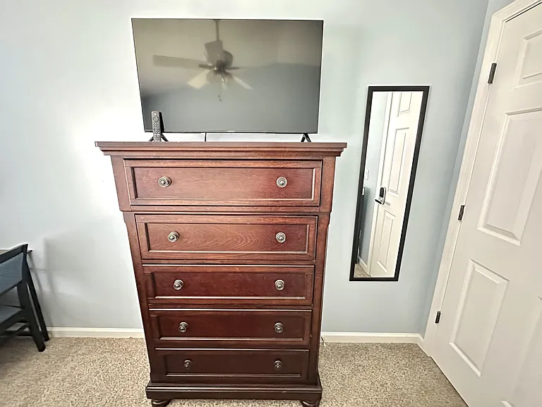 Furnished bedroom setup with a tall dresser, mounted TV, and full-length mirror for convenient storage and daily routines.