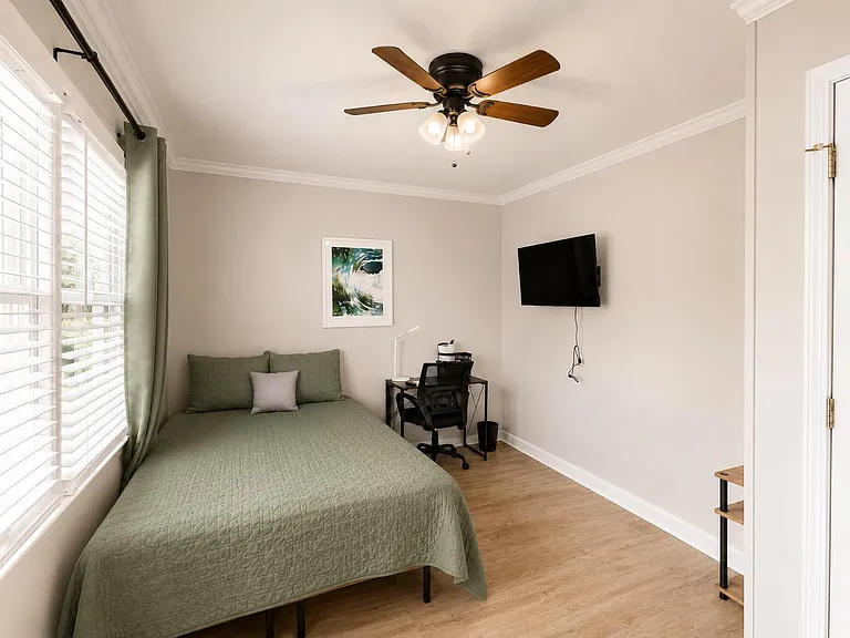 Private bedroom with a full bed, large window with blinds, ceiling fan, wall-mounted TV, and a compact work desk.