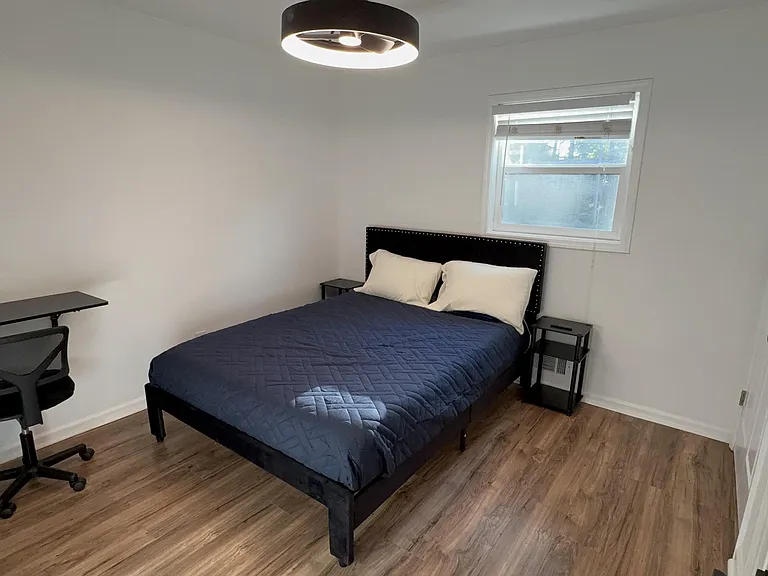 Furnished bedroom featuring a full bed, nightstands, and a dedicated desk area, plus a window and wood-look flooring.