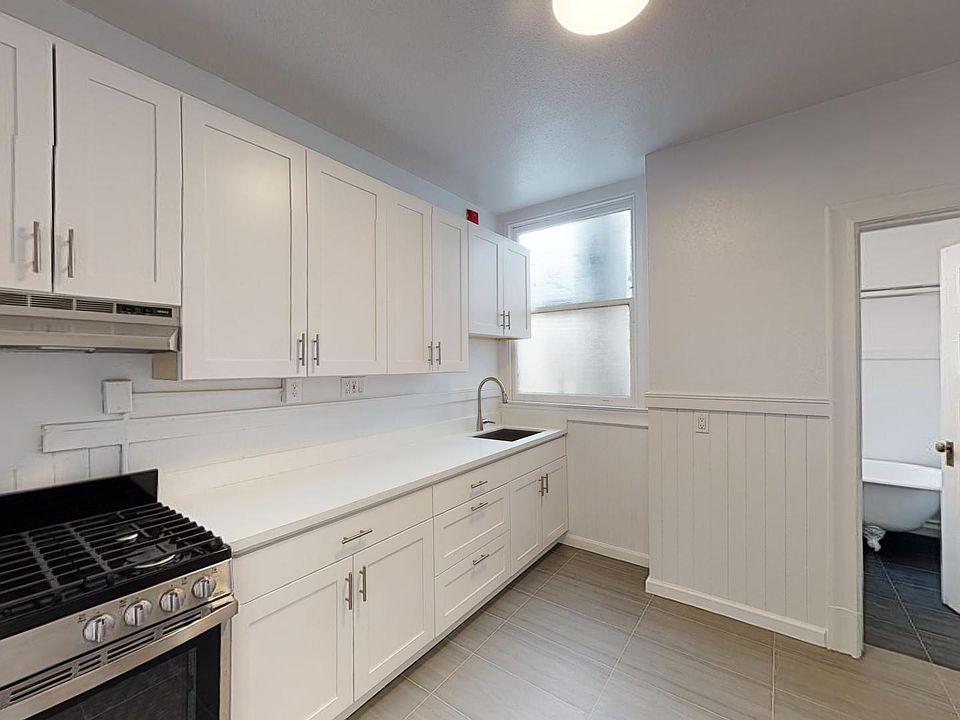 Kitchen with cabinets, stainless steel appliances, and a sink beneath a window, opening to a room with a bathroom