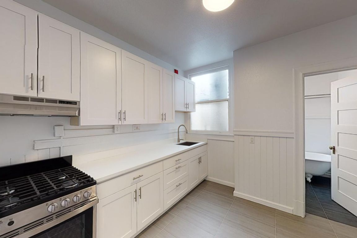 Kitchen with cabinets, stainless steel appliances, and a sink beneath a window, opening to a room with a bathroom