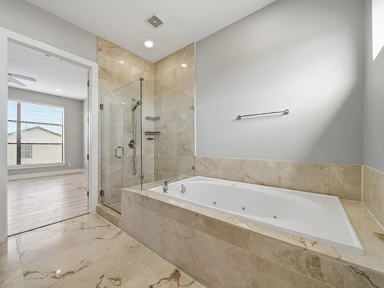Modern primary bathroom showcasing a sleek, upscale design with marble tile flooring and wall accents. A spacious glass-enclosed shower & built-in soaking tub. Seamlessly connected to a bright primary bedroom featuring warm wood flooring and large windows