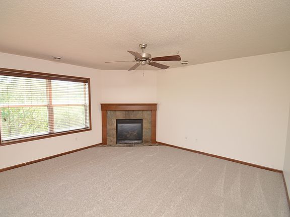 Bright and spacious freshly painted living area featuring new carpet, large windows for natural light, and a cozy gas fireplace with stone surround.