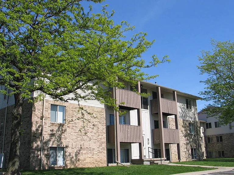 Brown brick and brown sided building with wooded balconies on the second and third floors. There is a large green tree to the left of the photo.