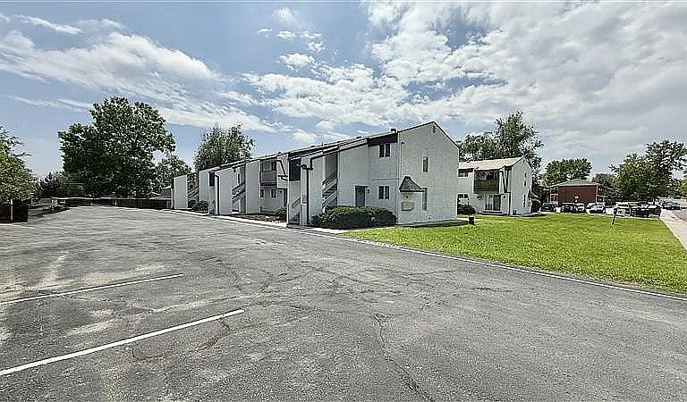 Charming white apartment complex with lush greenery and ample parking under a bright, blue sky.