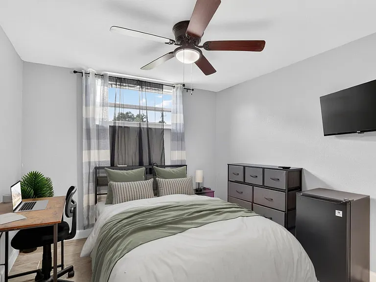 Furnished bedroom with a full bed, desk workspace, dresser storage, ceiling fan, and wall-mounted TV beside a sunny window.