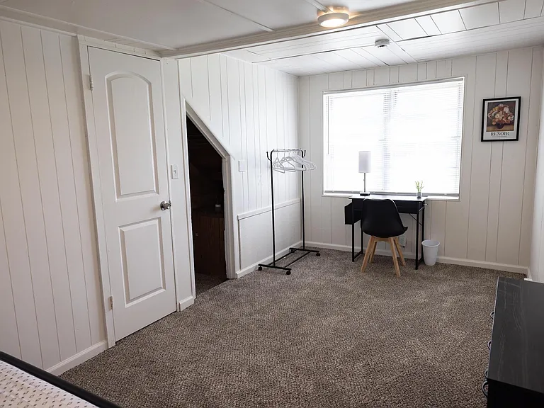 Carpeted bedroom with a large window, dedicated desk area, and convenient hanging rack for everyday storage.