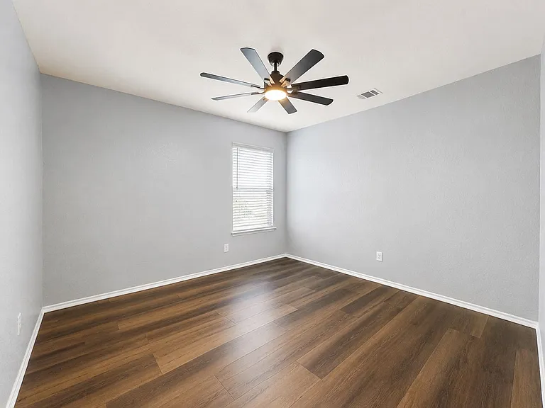 Spacious interior room featuring wood-finish flooring, light gray walls, a window with blinds, and a ceiling fan with integrated lighting