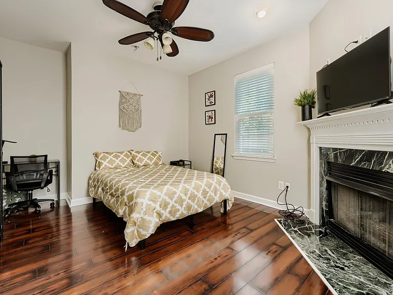 Spacious bedroom with hardwood floors, ceiling fan, desk area, and a decorative fireplace topped with a mounted TV.